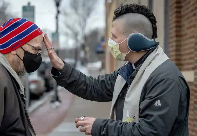 Clergy with mohawk places ashes on the forehead of a man with a winter cap. Both wear masks.