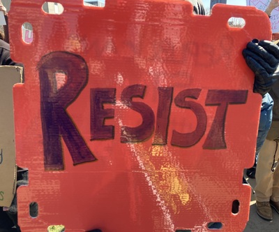 gloved hand holds handmade orange sign with bold dark letters saying RESIST